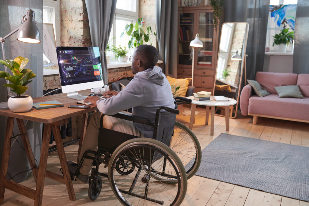 Rear view of African man sitting on wheelchair at the table in front of computer monitor and working with software at home