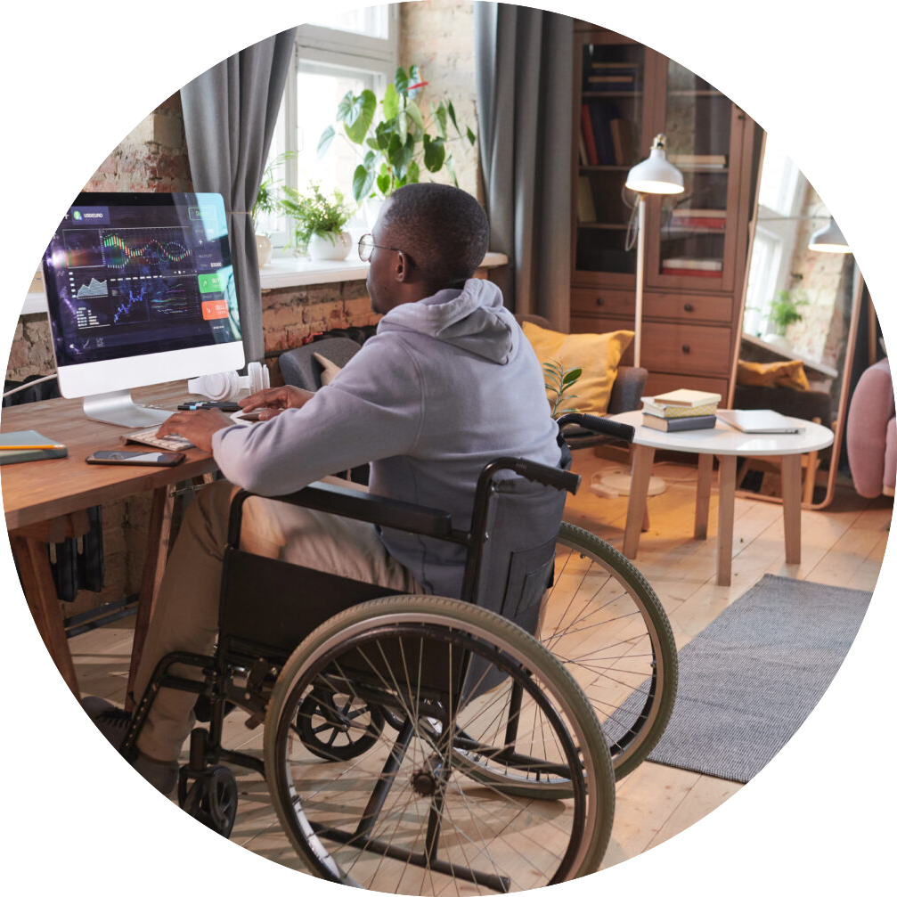 Rear view of African man sitting on wheelchair at the table in front of computer monitor and working with software at home